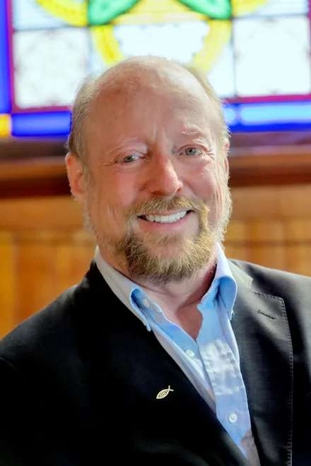 Rev. Dr. Tom Atkins, pastor of Rutland United Methodist Church, smiling in front of a stained-glass window